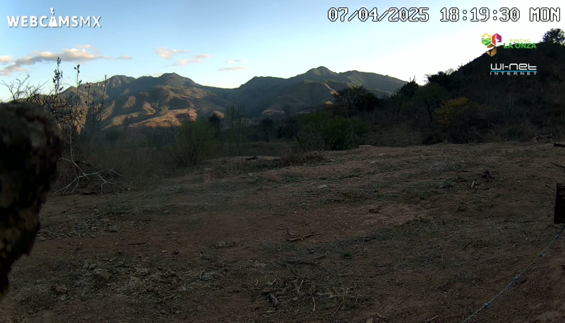 A dry, dirt clearing with sparse vegetation sits in front of a range of mountains under a clear, mostly sunny sky.