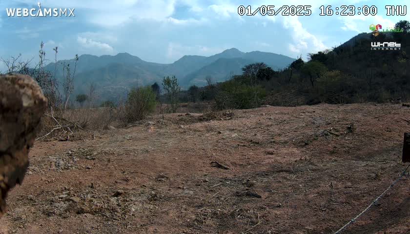 A mostly barren, reddish-brown landscape under a partly cloudy sky stretches towards a range of distant mountains.