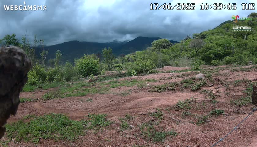 A dirt path winds through a sparsely vegetated, reddish-brown hillside under a cloudy sky, with mountains in the background.