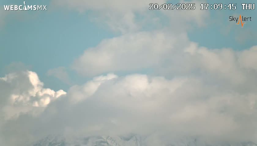 A mostly cloudy sky with puffy white clouds covers snow-capped mountains in the distance.