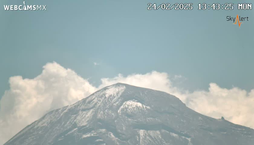 A snow-capped mountain is partially obscured by fluffy white clouds under a mostly clear, light-blue sky.
