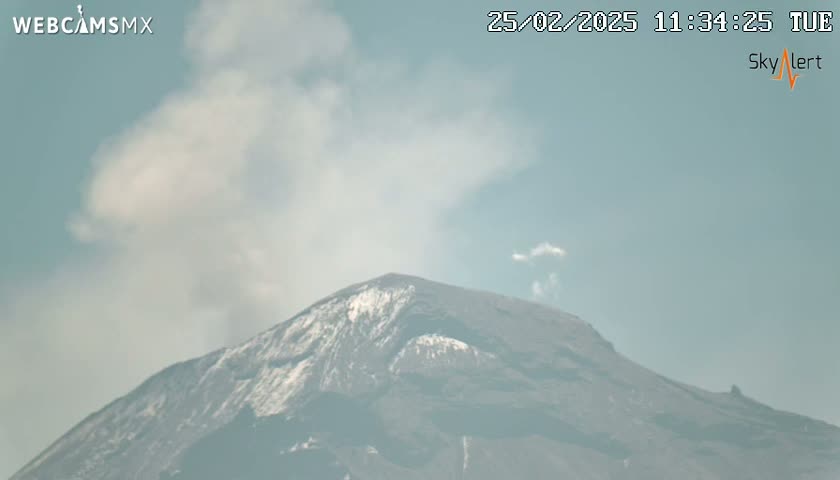 A partly cloudy sky overlooks a snow-capped mountain emitting a plume of smoke.