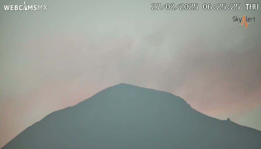 A hazy, pale pink sunrise illuminates a large, dark mountain.