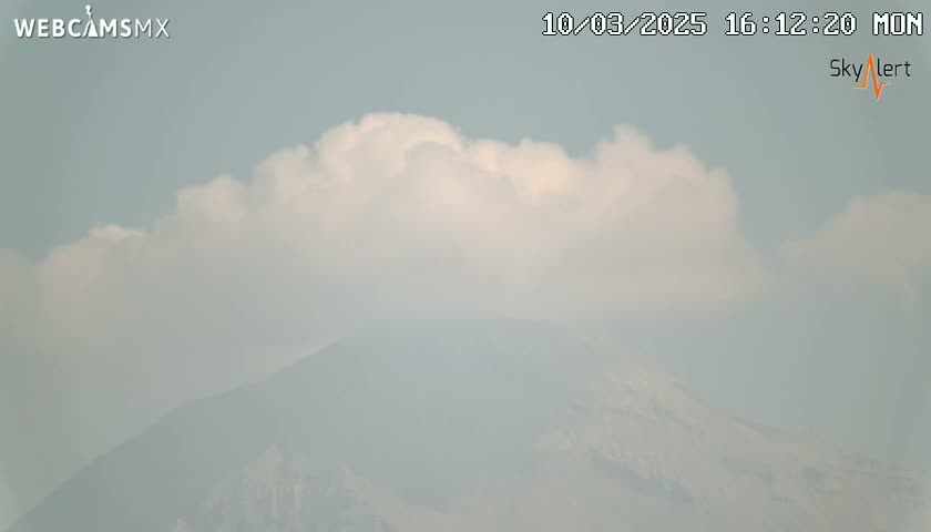 A hazy mountain is partially obscured by a layer of fluffy white clouds under a mostly clear sky.