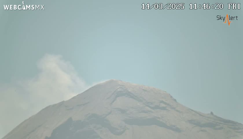 A large, dark-colored mountain is partially obscured by a light plume of smoke under a hazy, light-blue sky.