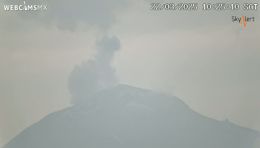 A hazy, overcast sky obscures a snow-capped mountain emitting a plume of smoke.