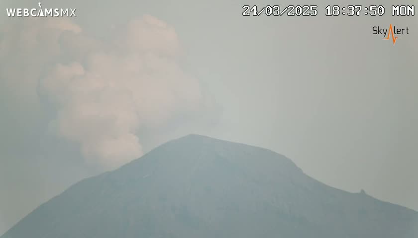 A hazy plume of smoke rises from a large, dark mountain under an overcast sky.