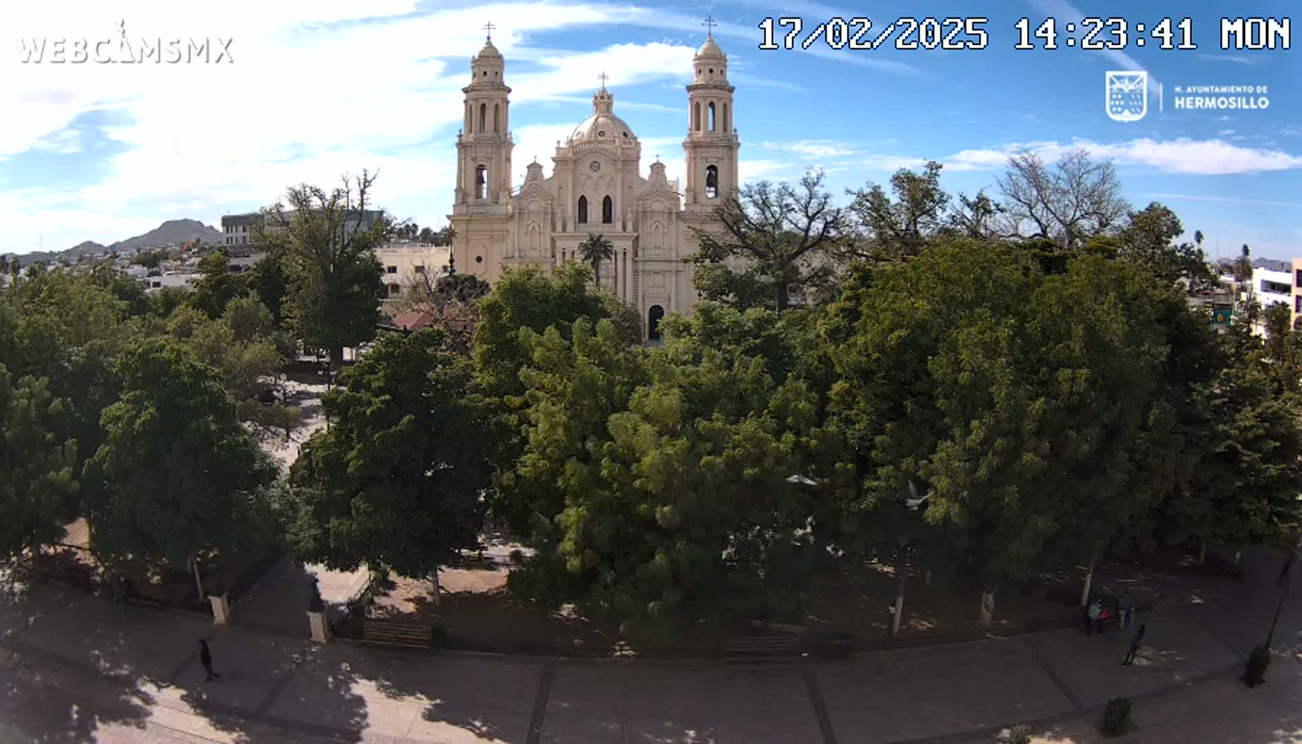 A large cathedral stands behind a plaza filled with leafy green trees under a mostly sunny sky.