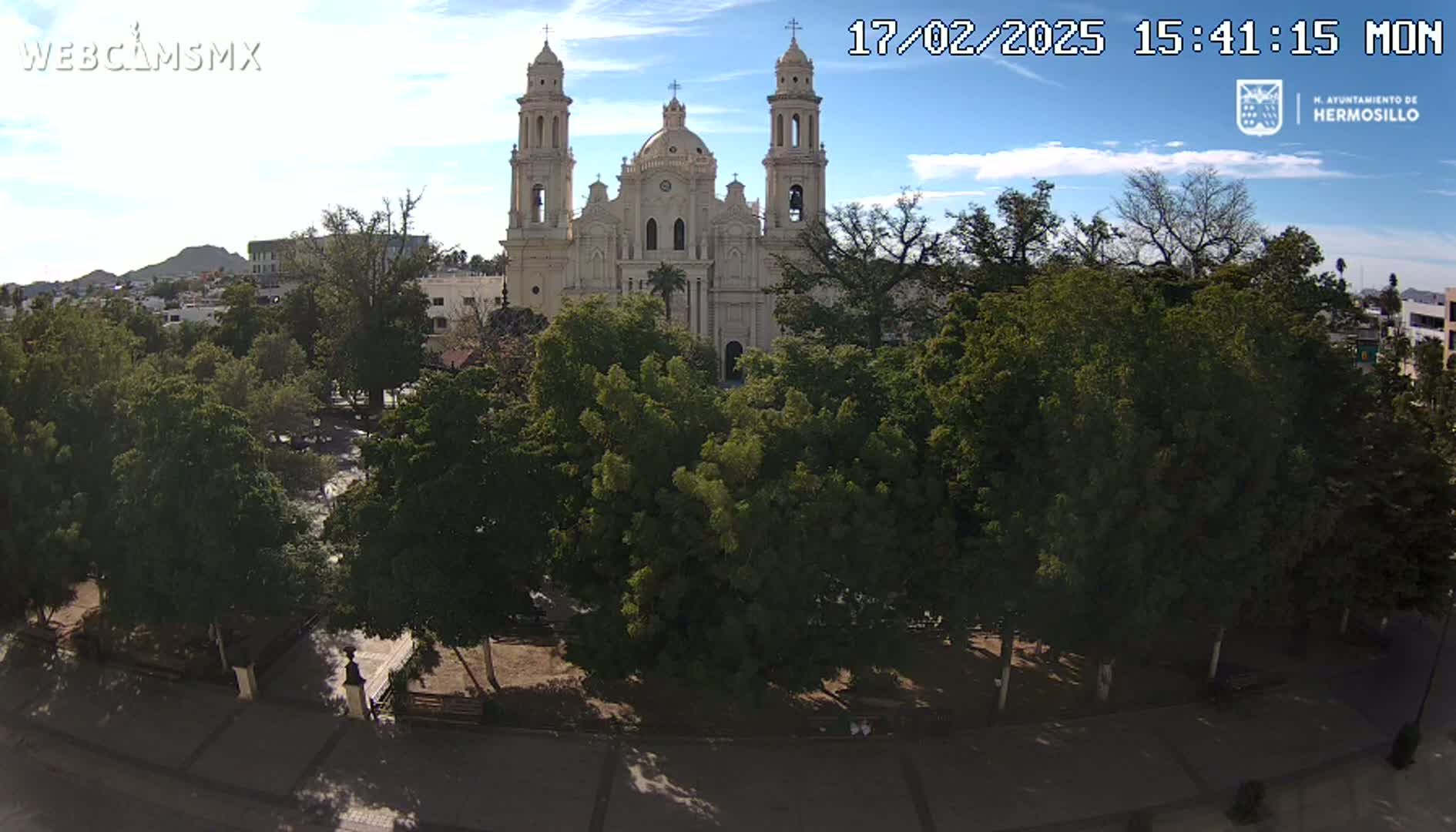 A large, light-colored cathedral is visible behind a park filled with lush green trees under a mostly sunny sky.