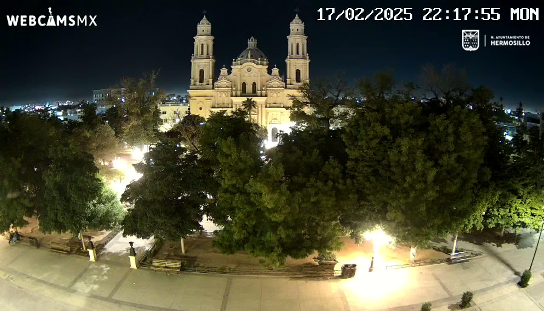 A large cathedral is visible at night, partially obscured by lush green trees in a plaza with walkways and benches.
