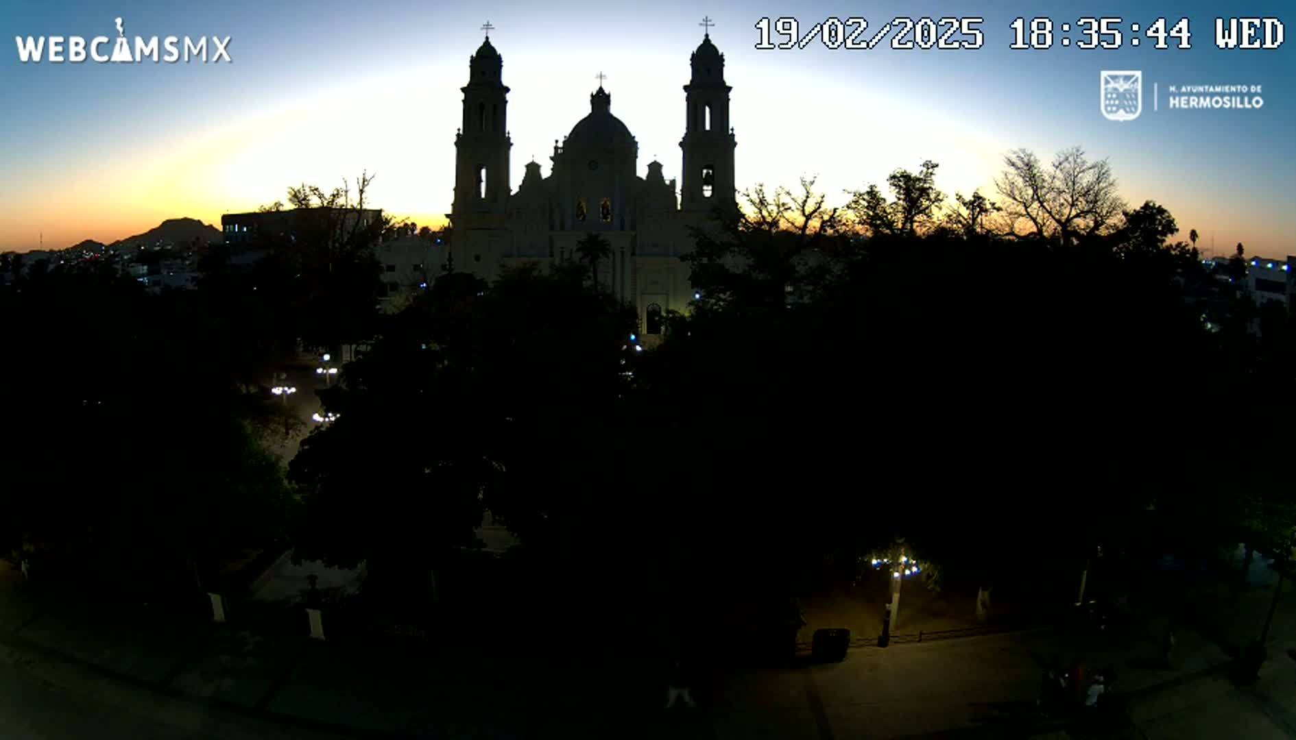 A large cathedral is silhouetted against a twilight sky, surrounded by dark trees and dimly lit pathways in a plaza.