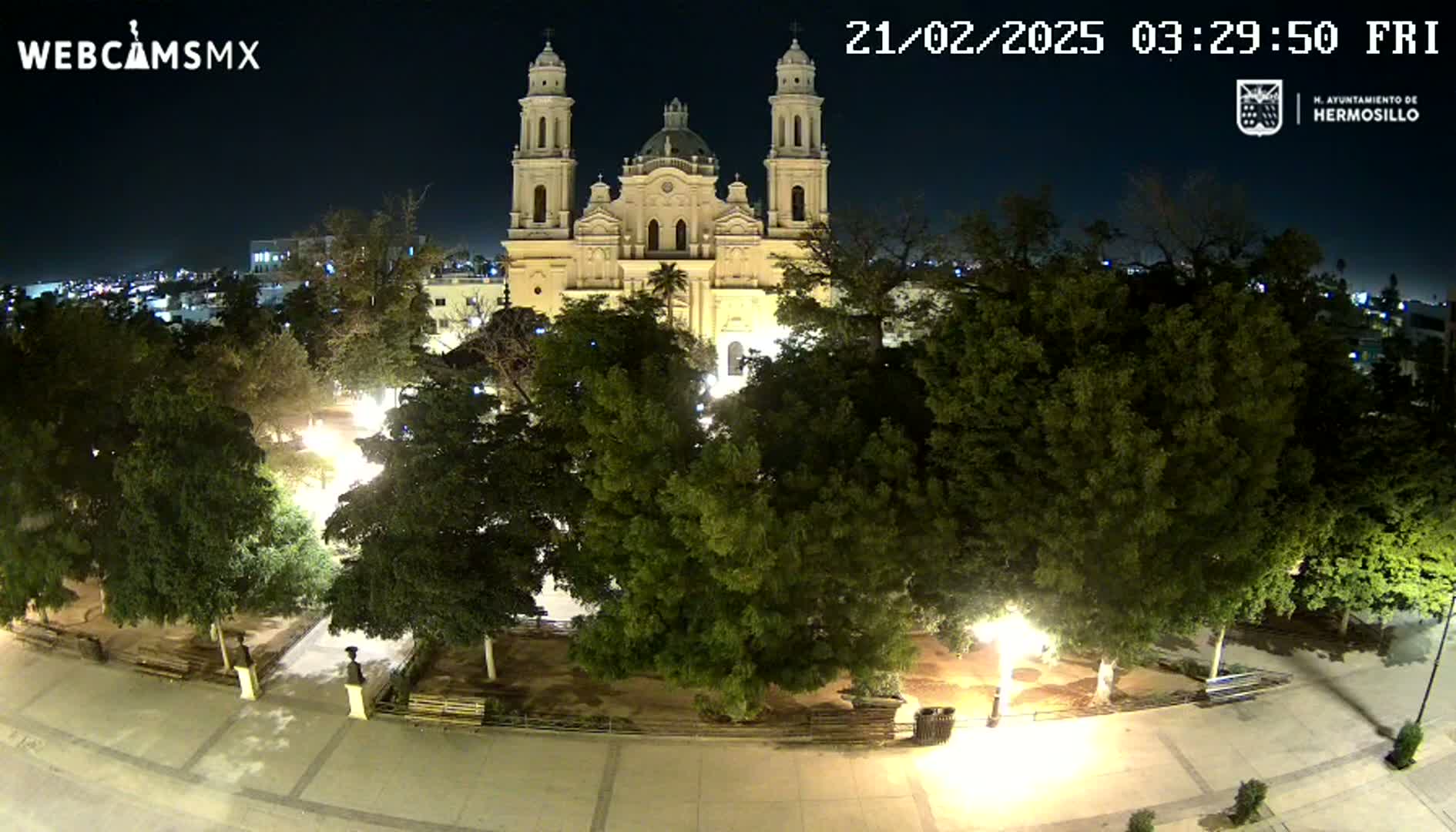 A brightly lit plaza at night, surrounded by leafy trees and centered on a large, pale stone cathedral.