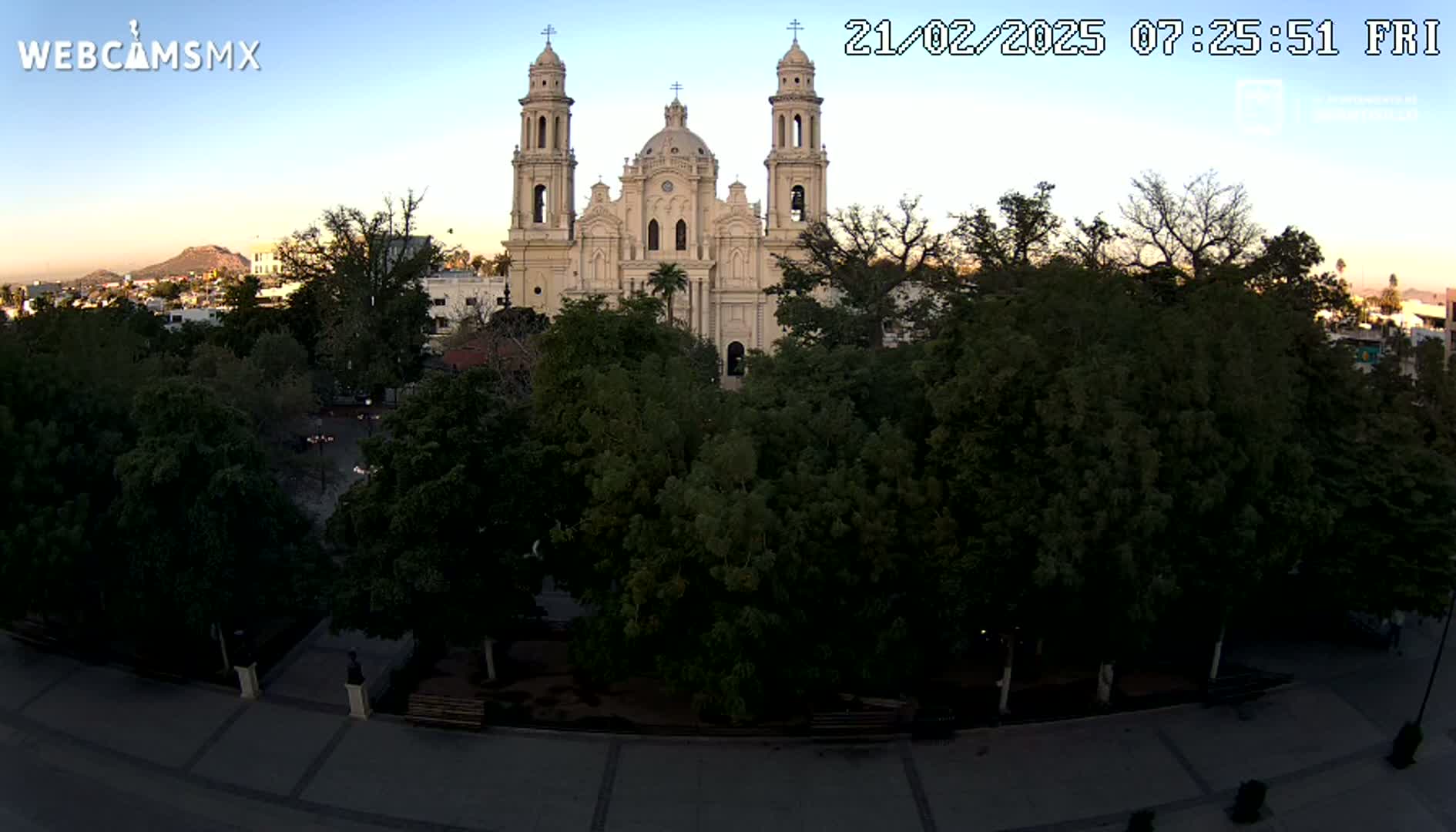 A large, light-beige cathedral stands behind a plaza filled with leafy green trees, under a clear, twilight sky.