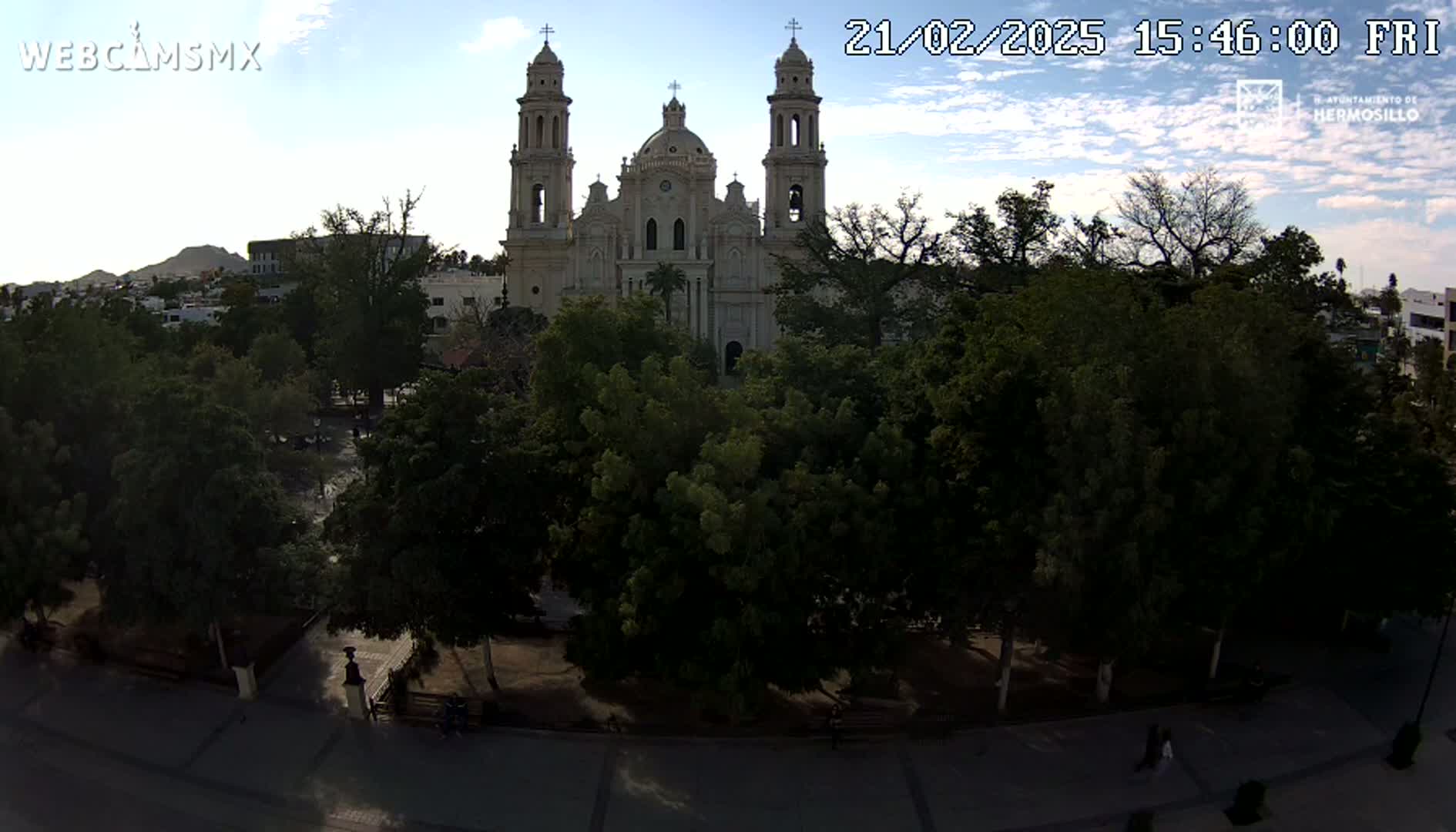 A large cathedral stands behind a plaza filled with lush green trees under a partly cloudy sky.