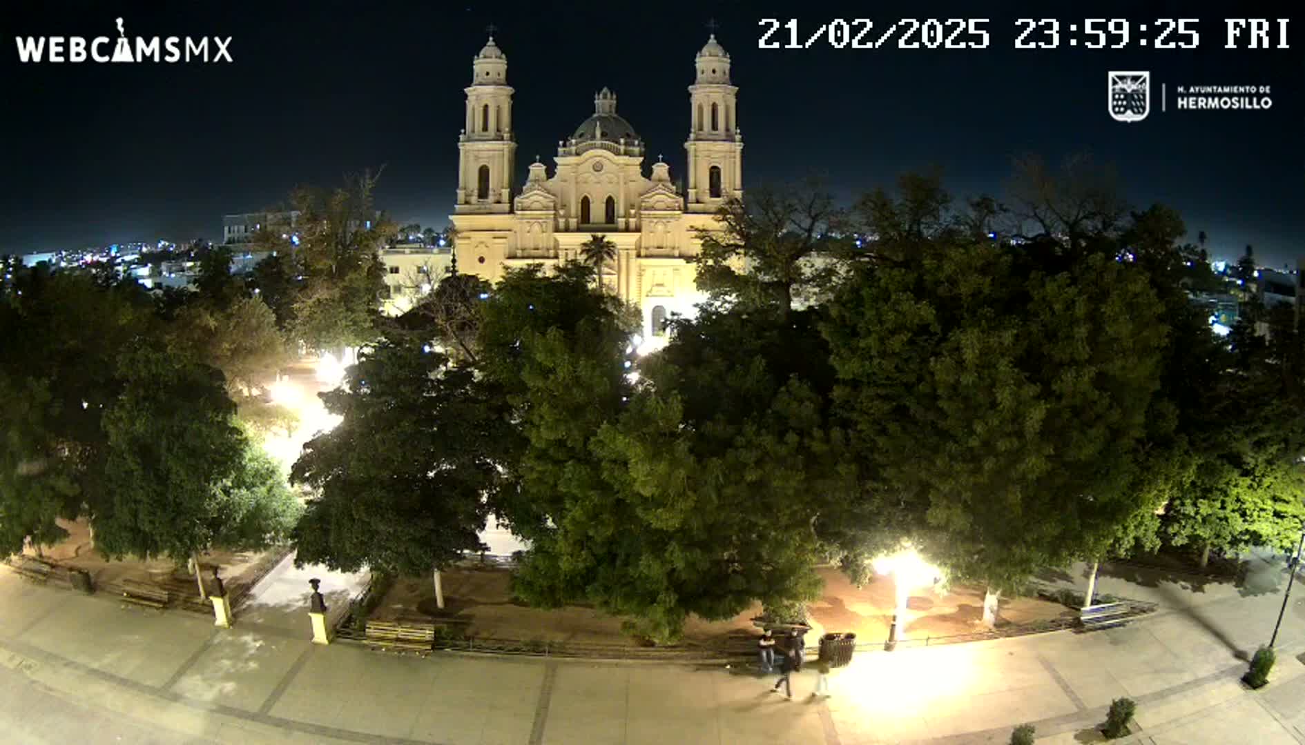 A brightly lit plaza at night, featuring a large cathedral in the background and numerous trees throughout.
