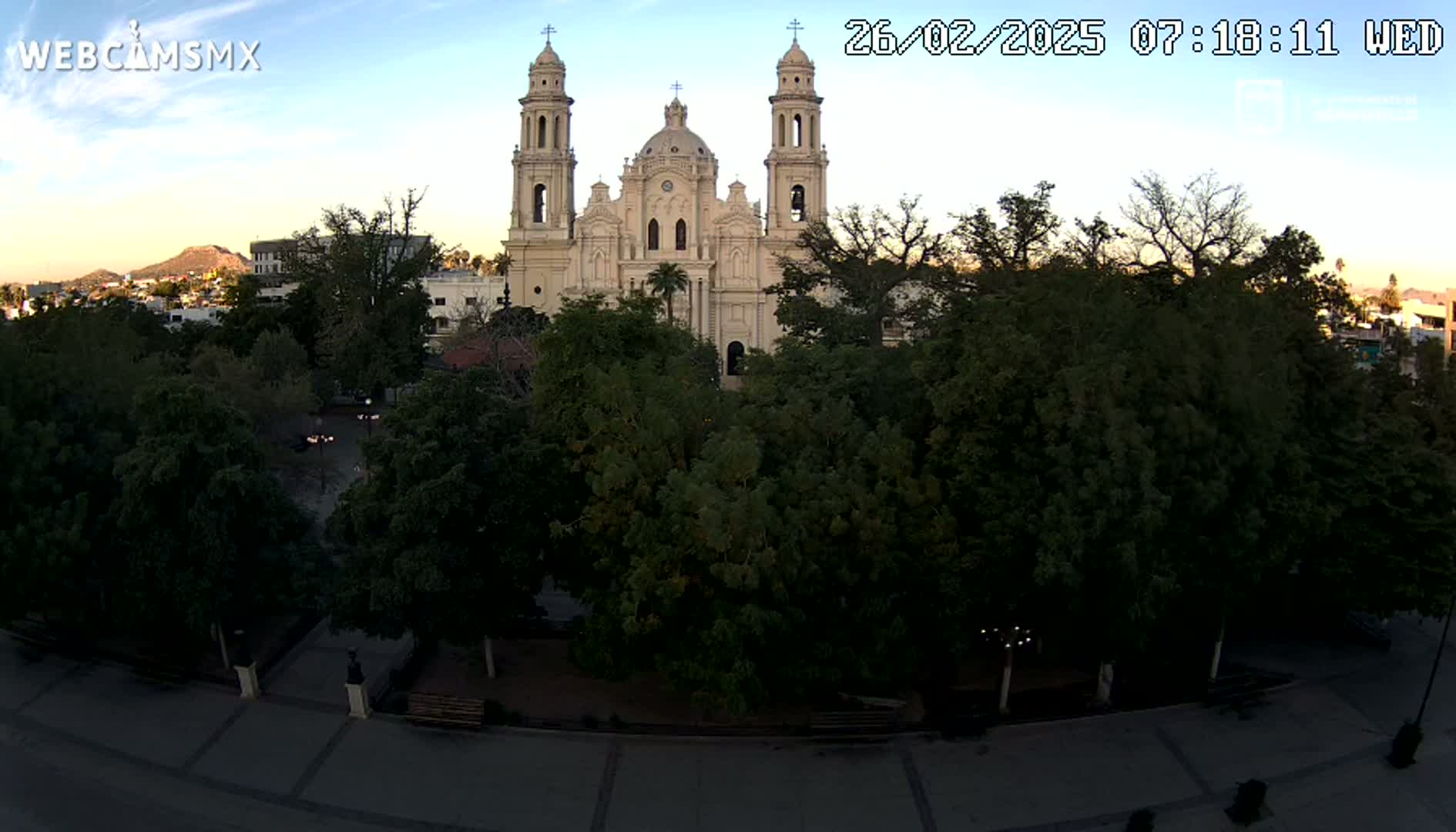 A large, light-colored cathedral stands behind a park filled with lush, green trees under a clear, twilight sky.