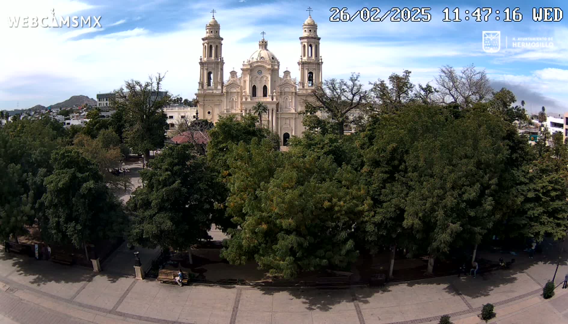 A large cathedral stands behind a plaza filled with leafy green trees under a partly cloudy sky.