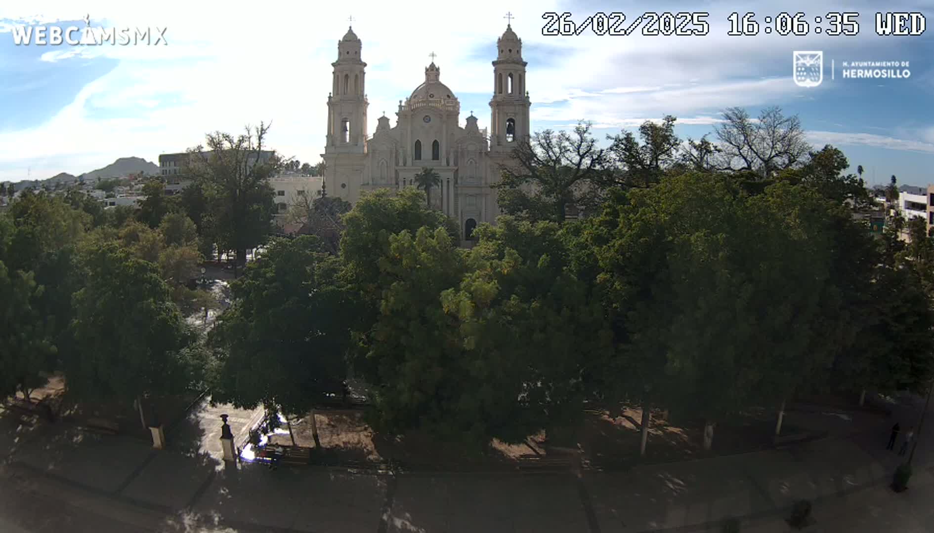 A large cathedral is partially obscured by lush green trees in a sunny plaza.