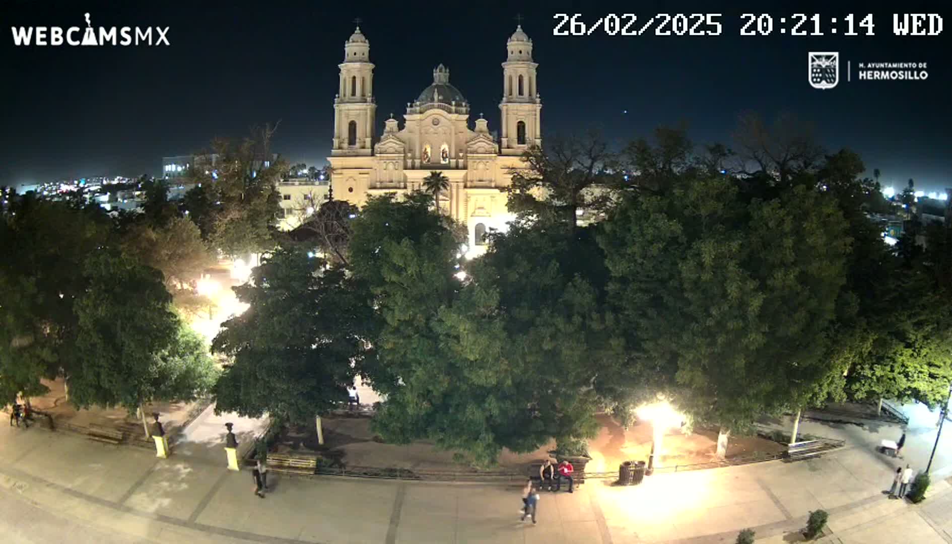 A brightly lit cathedral stands at the center of a plaza at night, surrounded by lush green trees and a few people walking around under a clear sky.