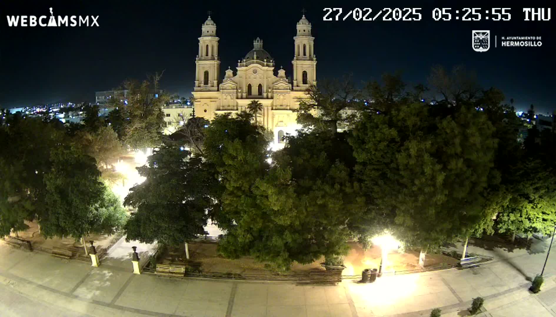 A large cathedral is seen at night, illuminated, surrounded by a plaza with numerous trees and lights.