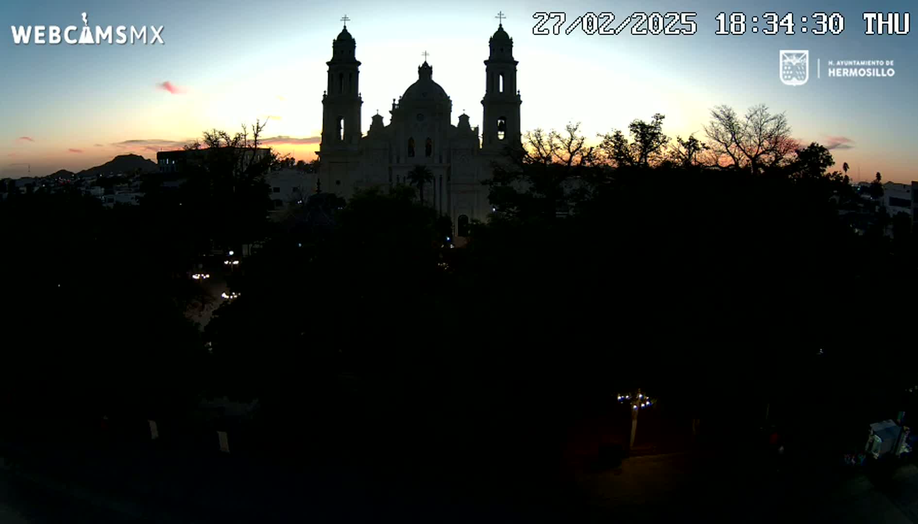 A large, light-colored cathedral is silhouetted against a twilight sky, with dark trees and a few dimly lit vehicles in the foreground.