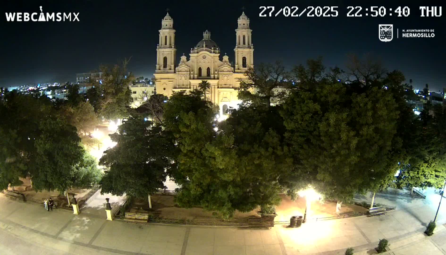 A brightly lit plaza at night, with a large cathedral in the background, surrounded by lush green trees under a clear, dark sky.
