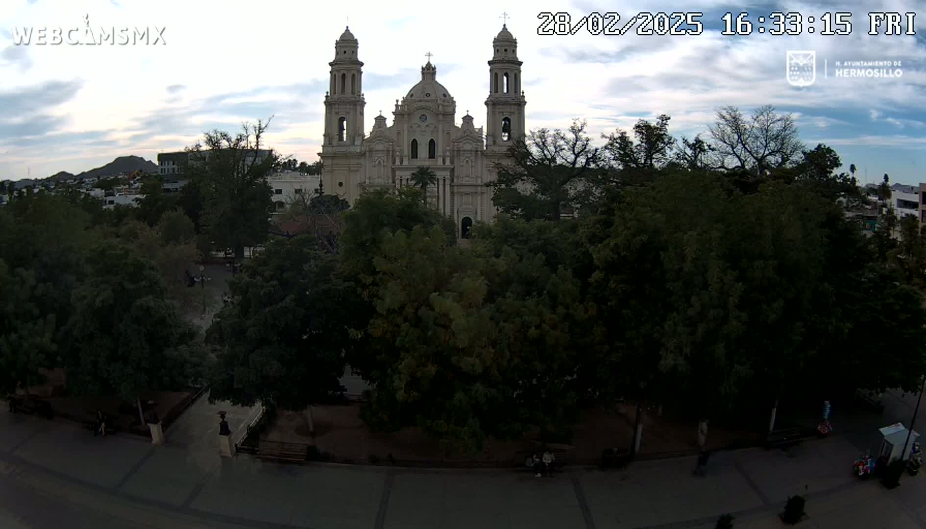 A large, light-colored cathedral stands behind a plaza filled with leafy green trees under a partly cloudy sky.