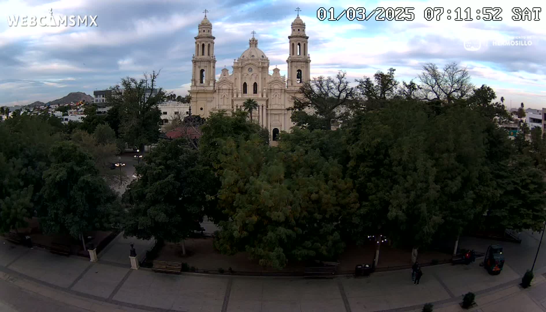 A large, light-colored cathedral is situated behind a park filled with lush green trees under a partly cloudy sky.