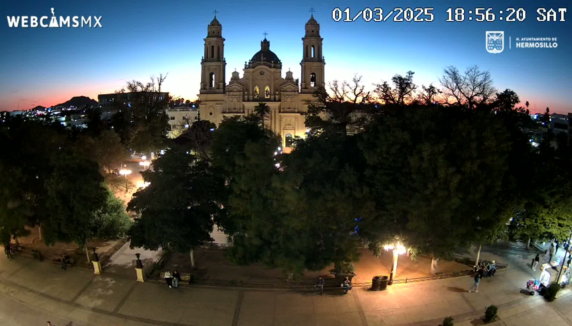 A large cathedral is illuminated at twilight, nestled within a tree-lined plaza where people are walking around.