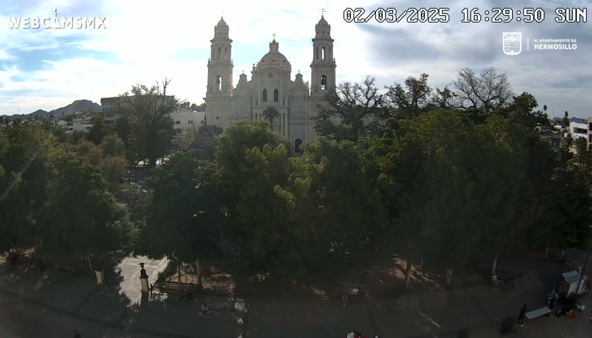 A large cathedral stands behind a leafy park under a partly cloudy sky.