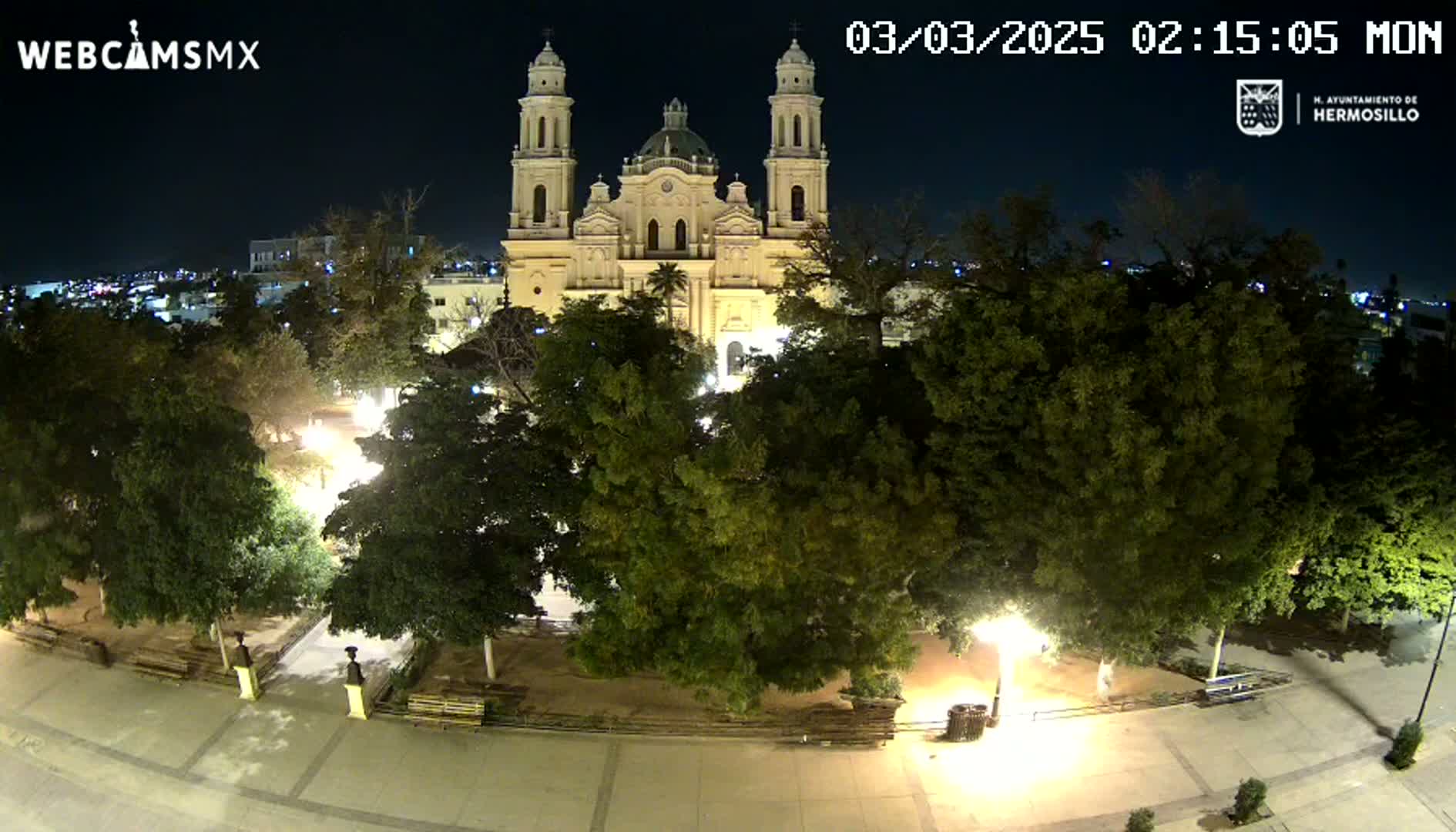 A brightly lit plaza at night, centered around a large cathedral, surrounded by lush green trees under a clear, dark sky.