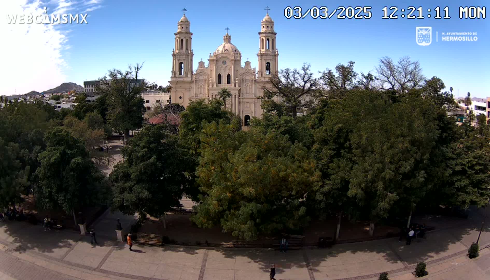 A large cathedral is visible behind a plaza filled with lush green trees under a clear blue sky.
