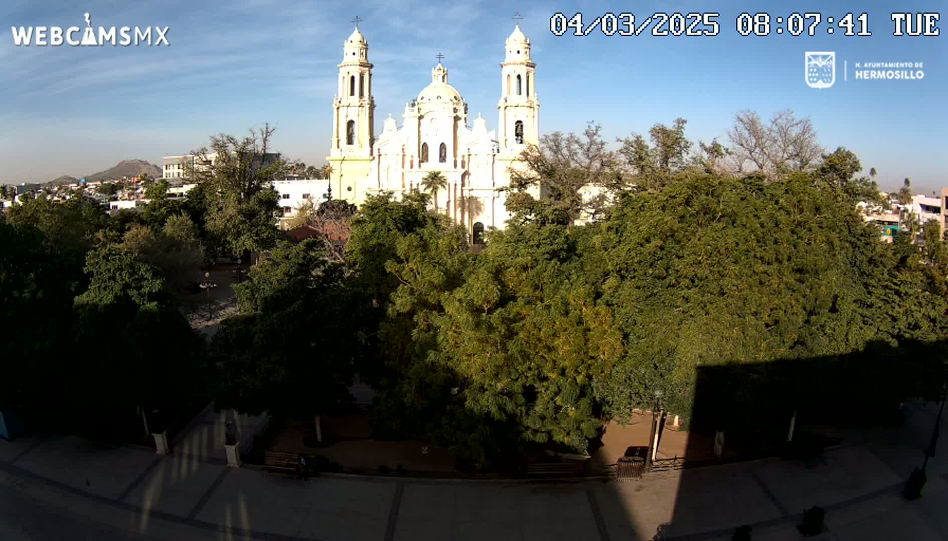 A large white cathedral is visible beyond a plaza filled with lush green trees under a clear, sunny sky.
