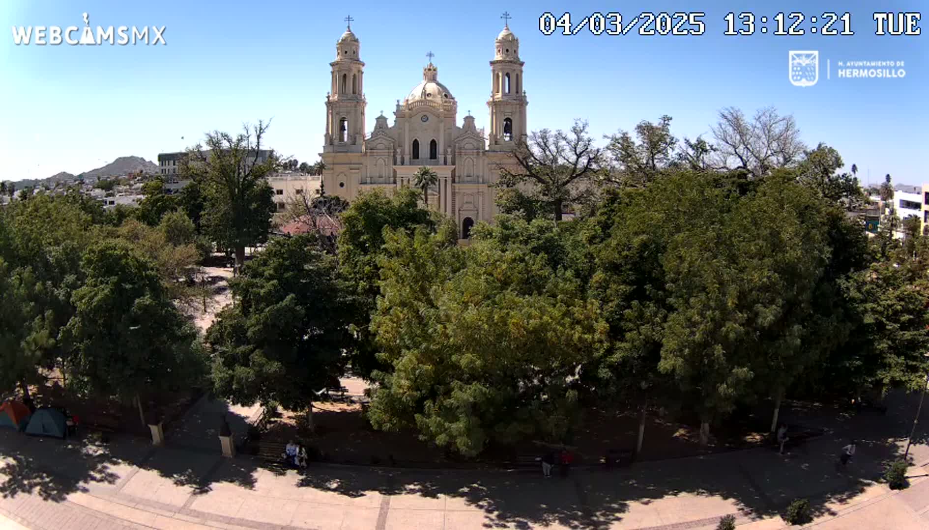 A large tan cathedral stands behind a lush green park on a sunny day.