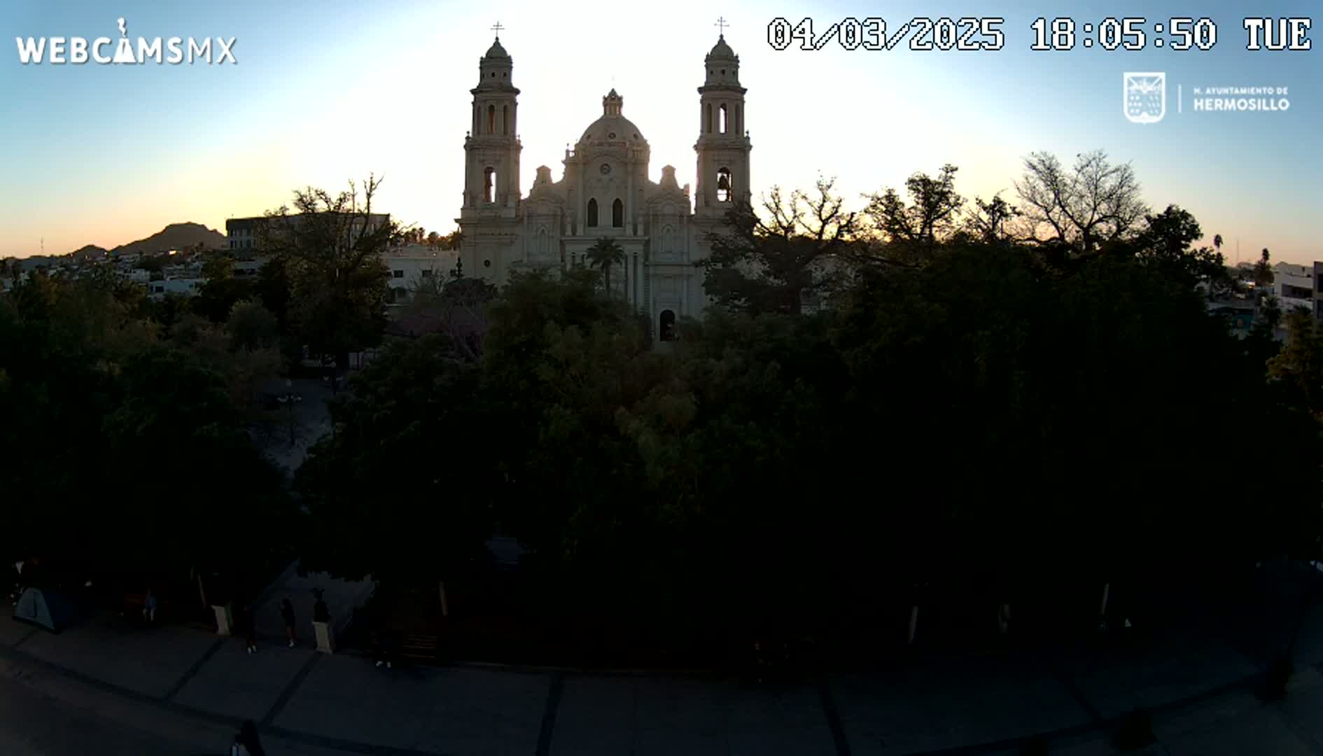 A large, light-colored cathedral stands in the center of a plaza surrounded by dark, leafy trees at dusk under a clear sky.