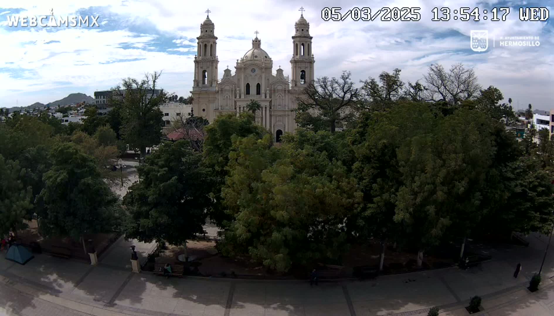A large cathedral stands behind a plaza filled with leafy green trees under a partly cloudy sky.