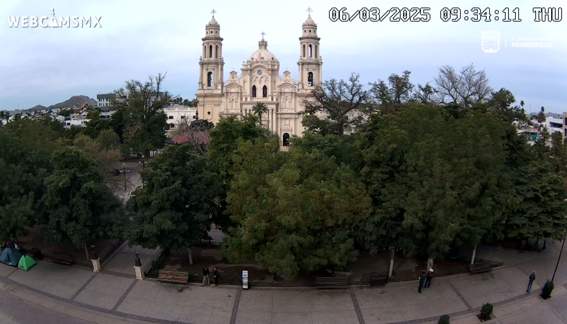 A large cathedral stands behind a plaza filled with lush green trees under a mostly cloudy sky.