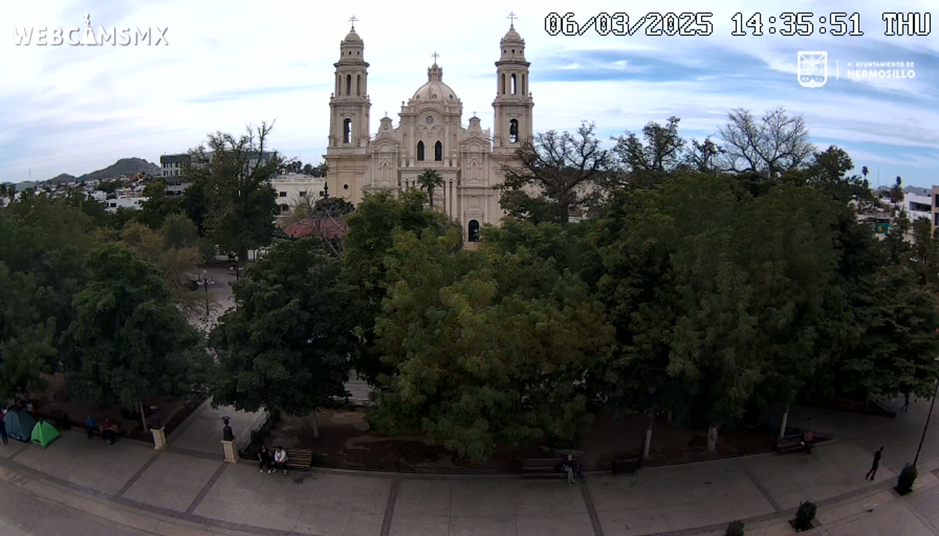 A large cathedral stands behind a plaza filled with leafy green trees under a partly cloudy sky.