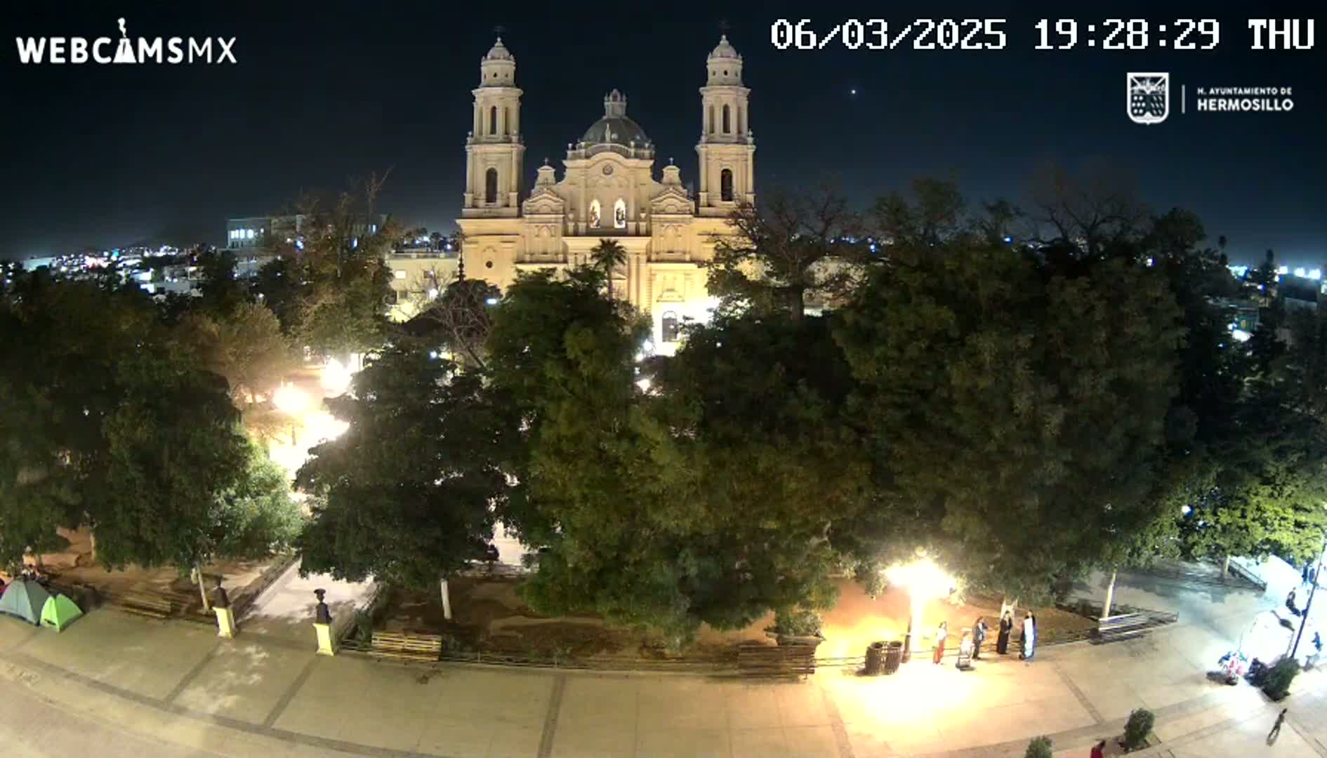 A brightly lit plaza at night, surrounded by trees, with a large cathedral in the background under a clear, dark sky.