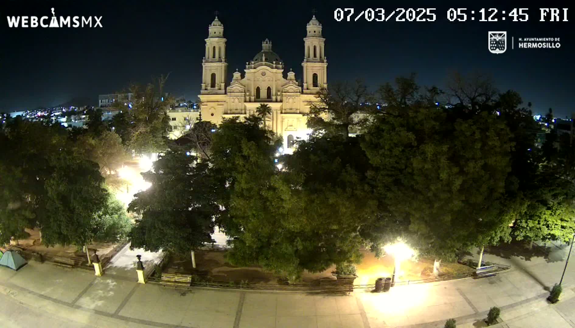 A large, illuminated cathedral stands at night behind a plaza filled with trees and illuminated walkways.