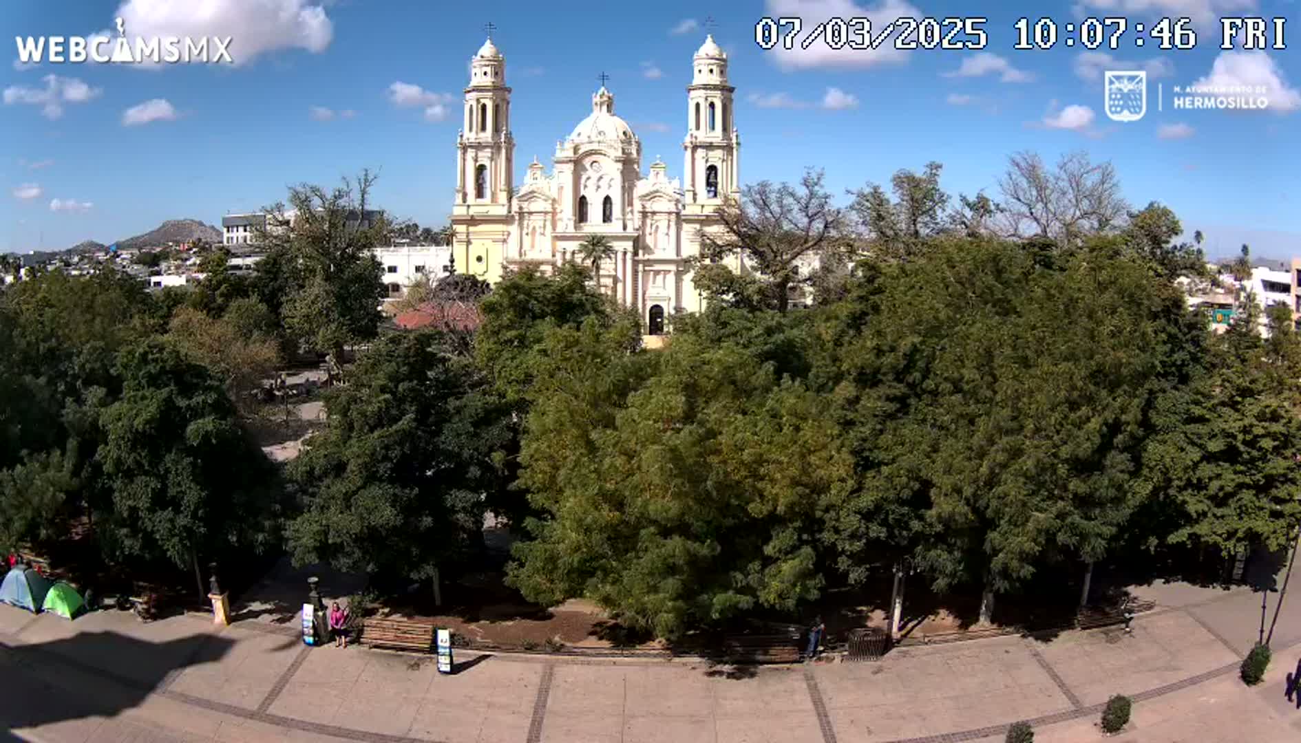A large, light-yellow cathedral stands behind a plaza filled with lush green trees under a mostly sunny sky.