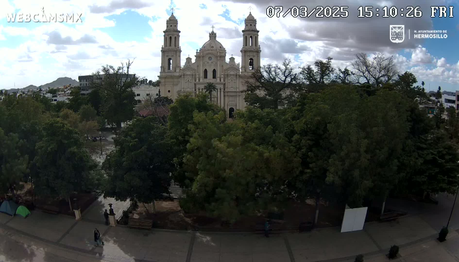 A large white cathedral is visible behind a plaza filled with lush green trees under a partly cloudy sky.