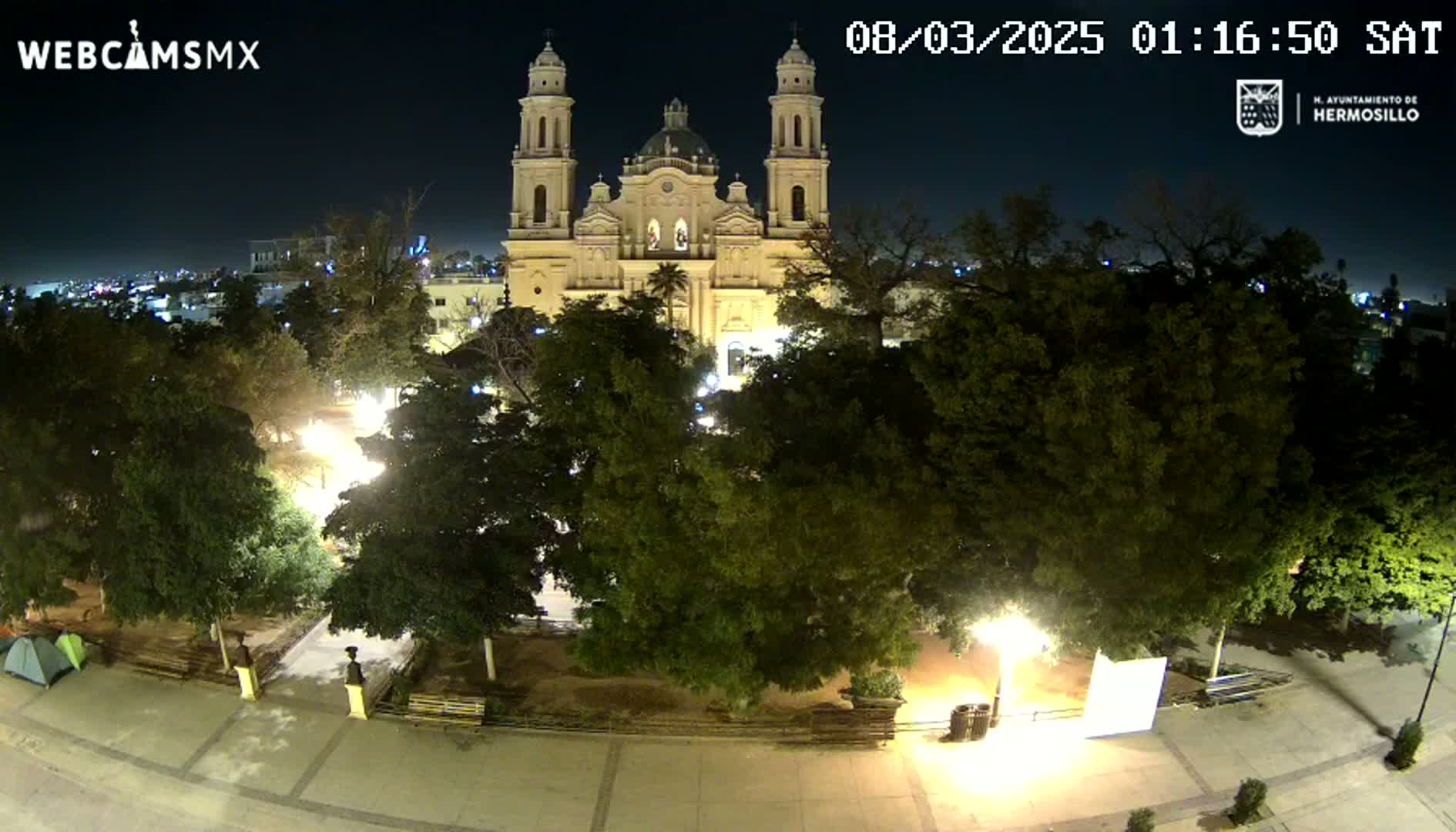 A nighttime, high-angle view shows a large cathedral illuminated, surrounded by a plaza with trees and park benches under a clear sky.