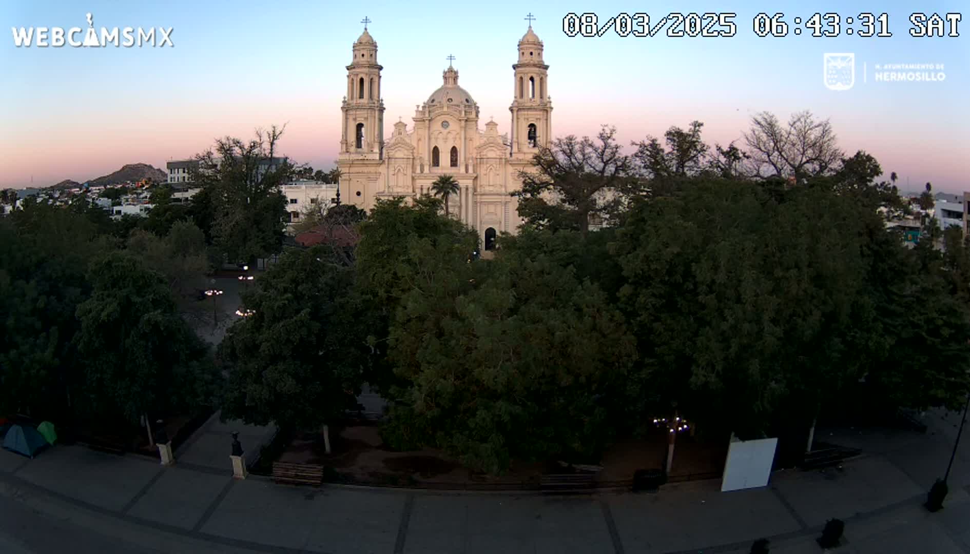 A large, light-colored cathedral stands behind a square filled with lush green trees under a twilight sky.