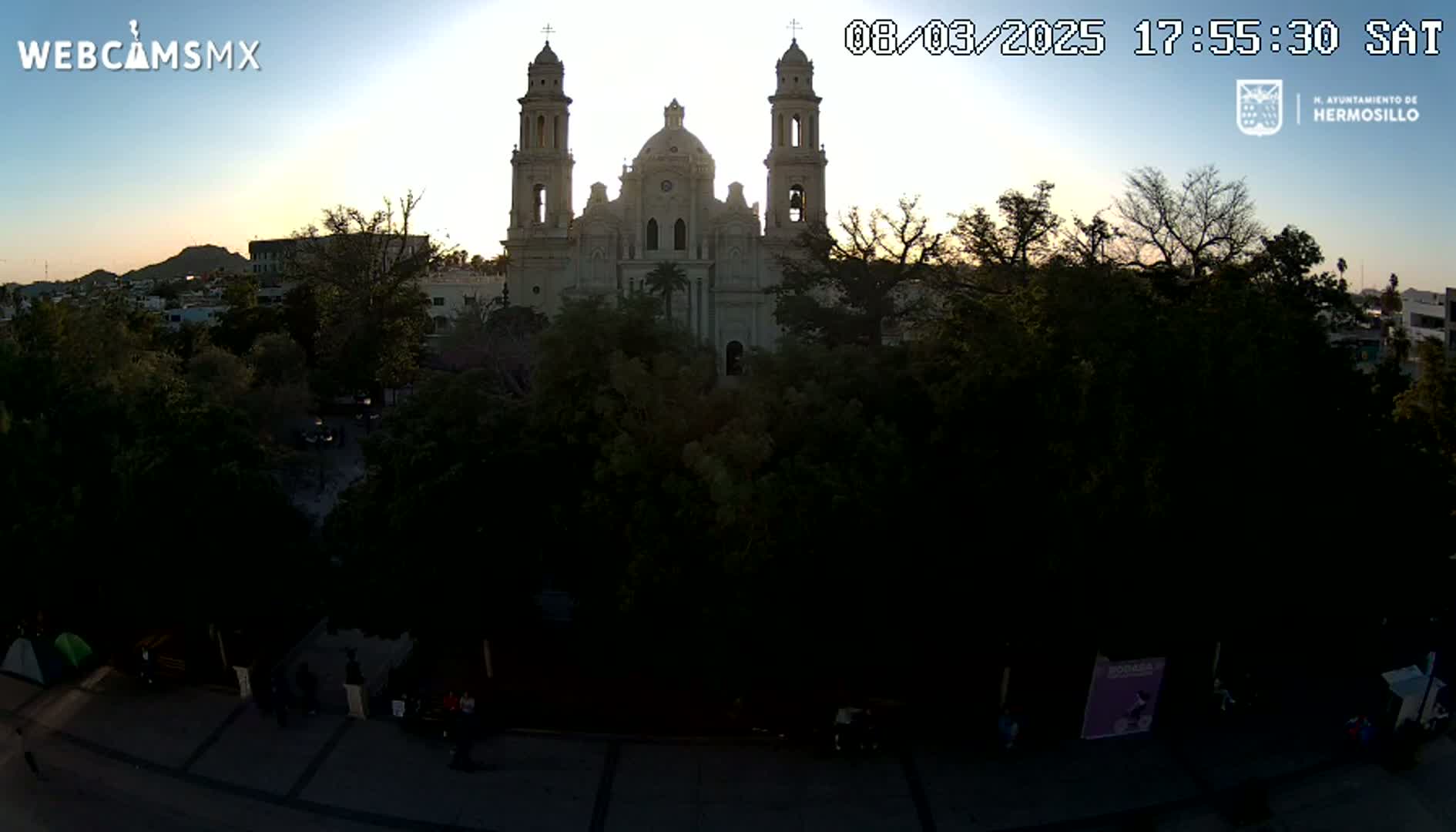 A large cathedral stands in the center of a town square surrounded by dark green trees at dusk under a clear sky.