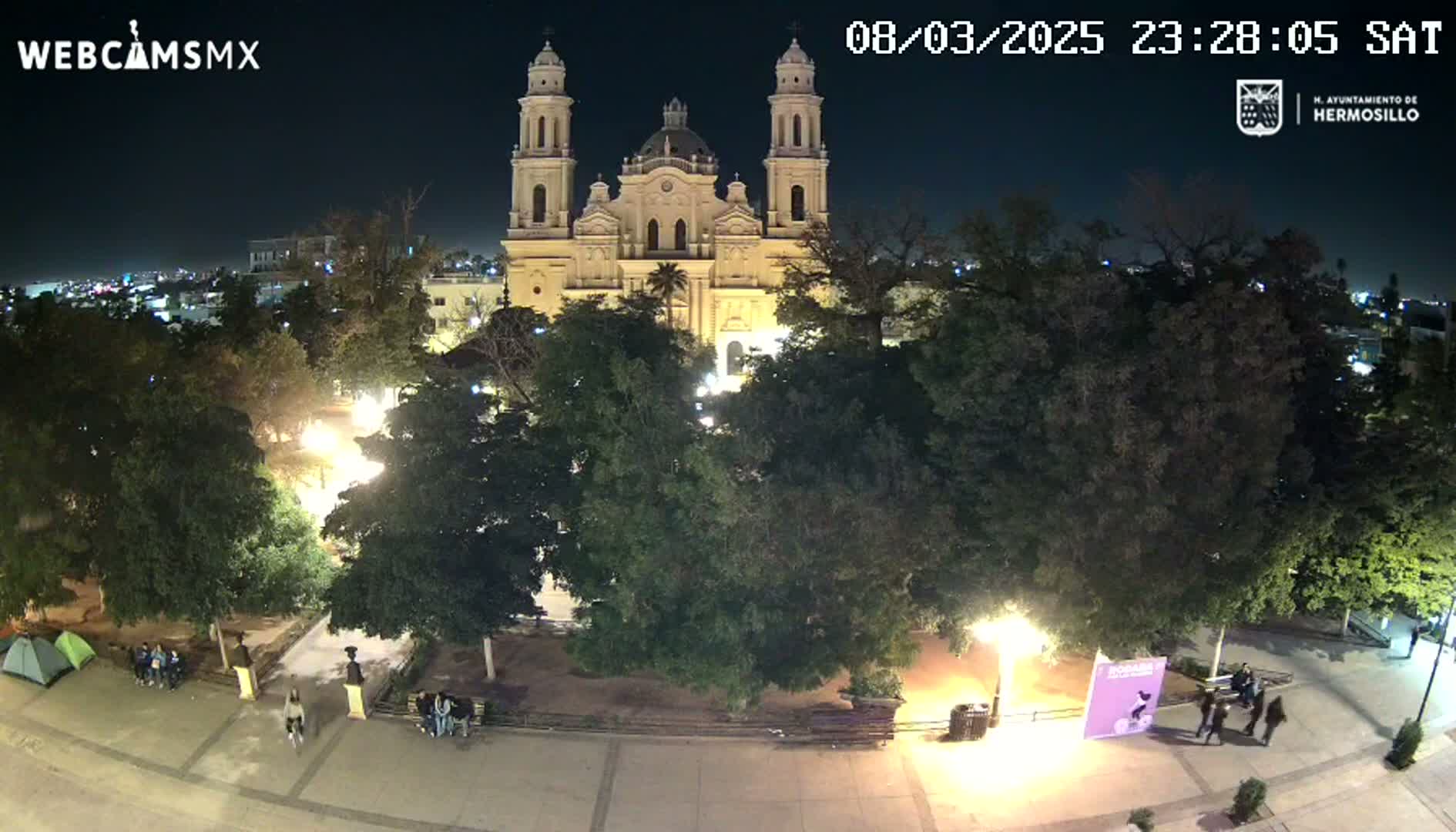 A nighttime aerial view shows a large cathedral surrounded by a plaza with trees and a few people, under a clear sky.