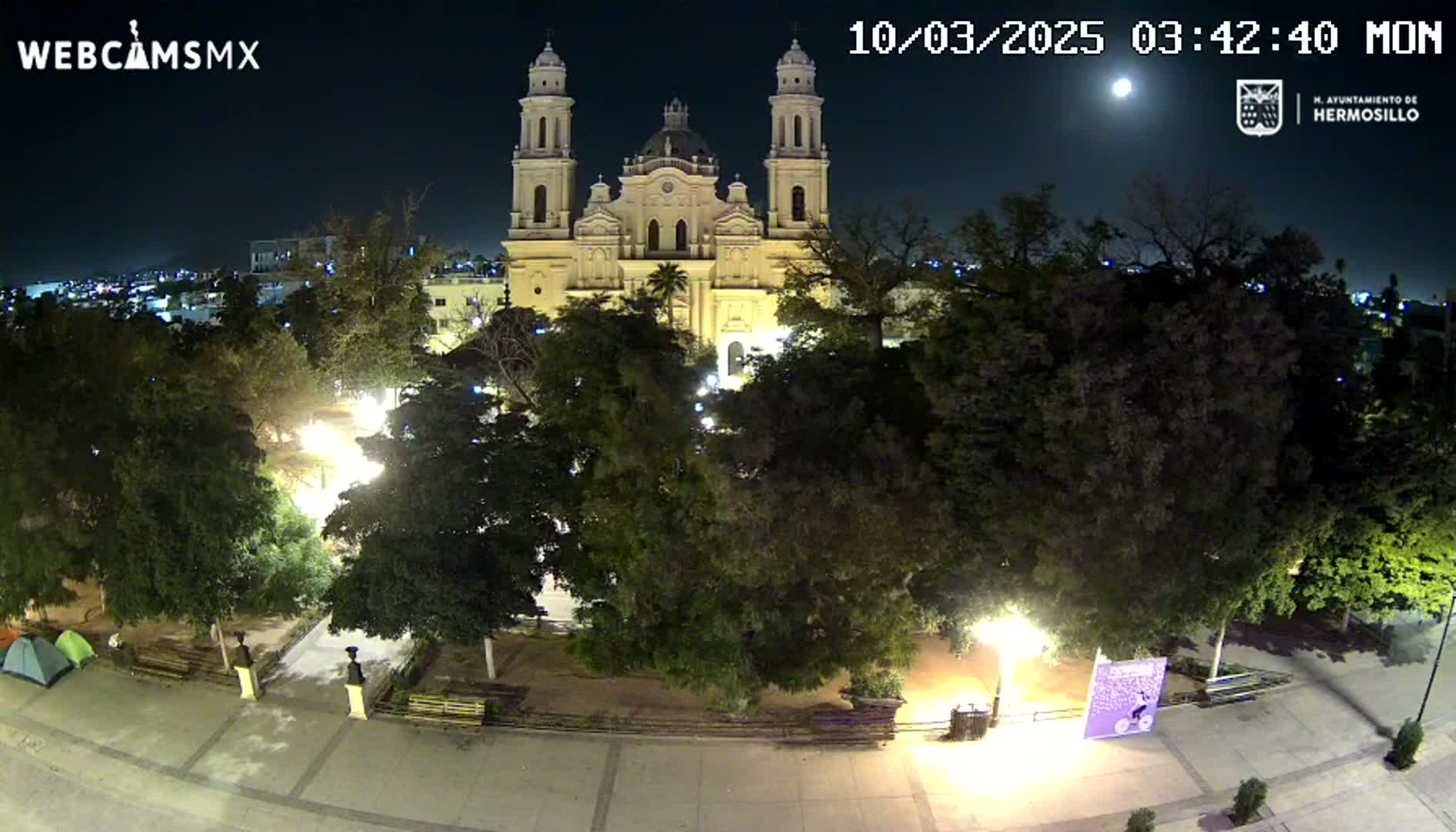 A brightly lit plaza at night, featuring a large cathedral in the background under a full moon.