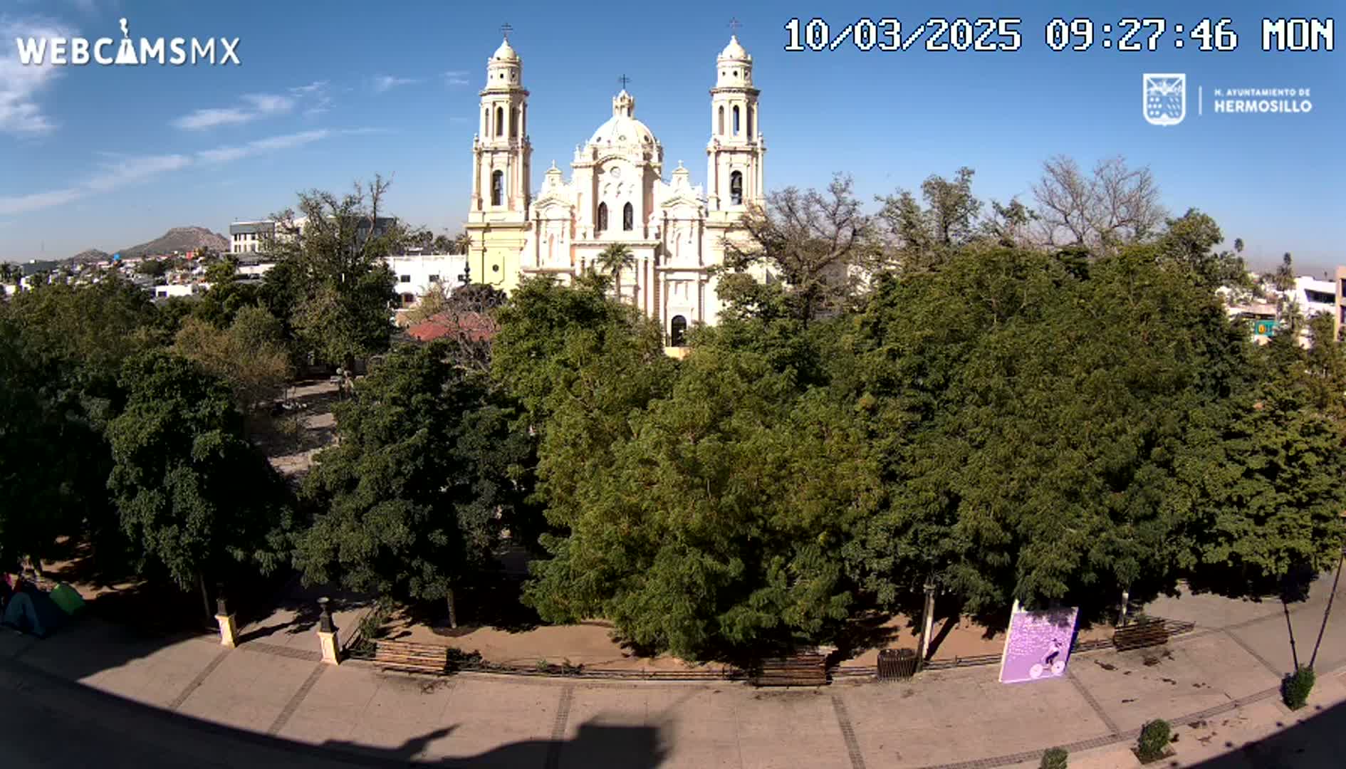 A large, light-colored cathedral stands behind a park filled with lush green trees under a clear, sunny sky.
