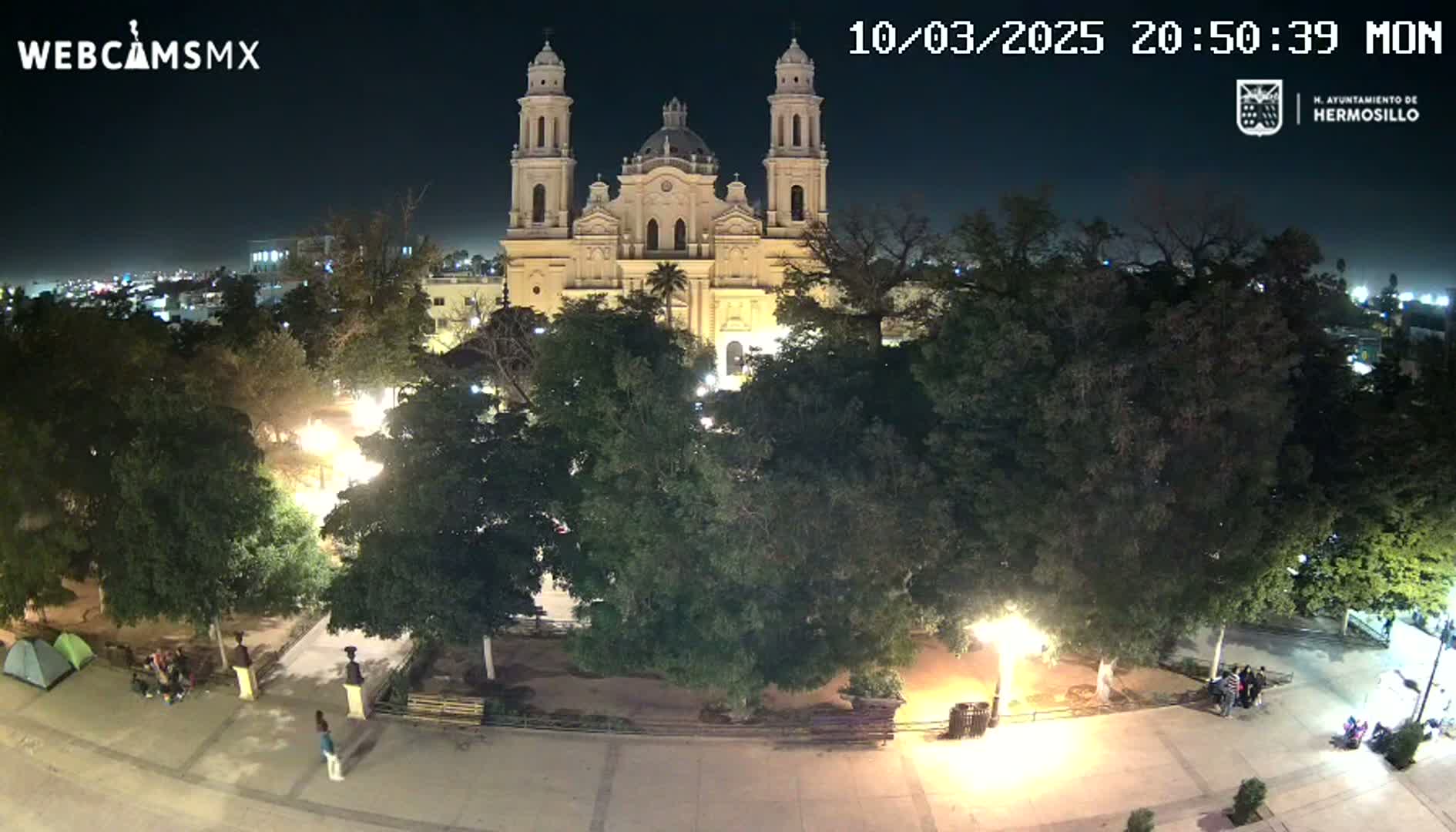 A nighttime, overhead view of a plaza with a large cathedral at its center, surrounded by lush green trees and a few people.