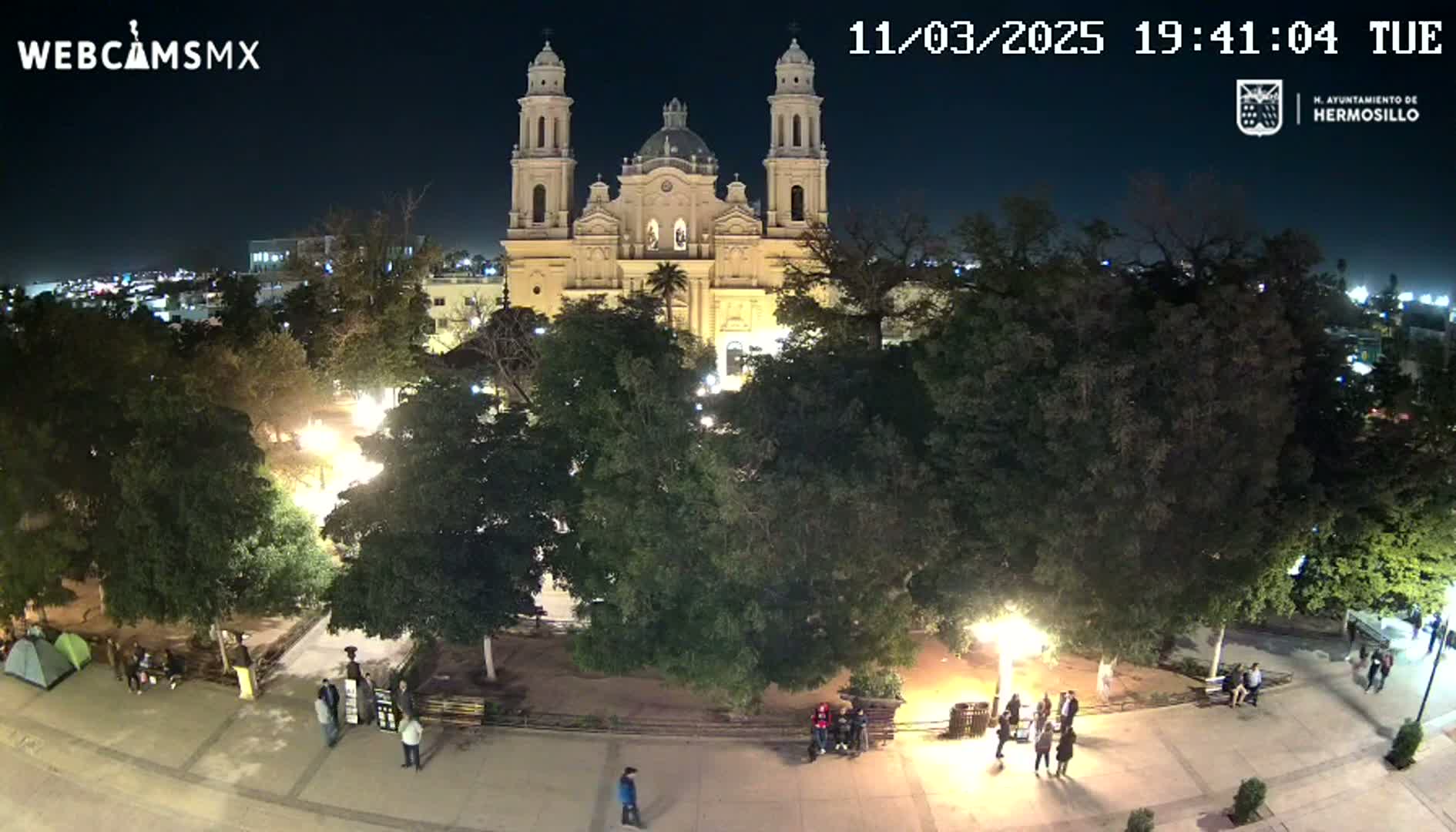 An illuminated cathedral stands at night in a park surrounded by trees and a few people, under a clear, dark sky.